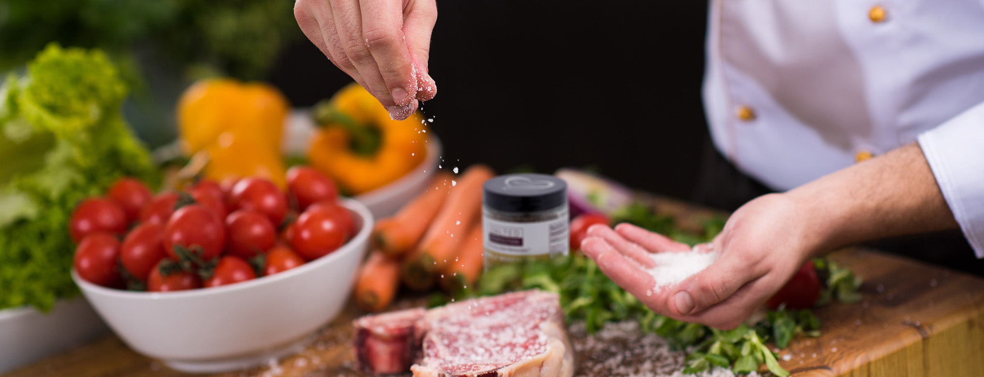 Chef sprinkling Simply Rosemary salt onto a steak with fresh vegetables in the background